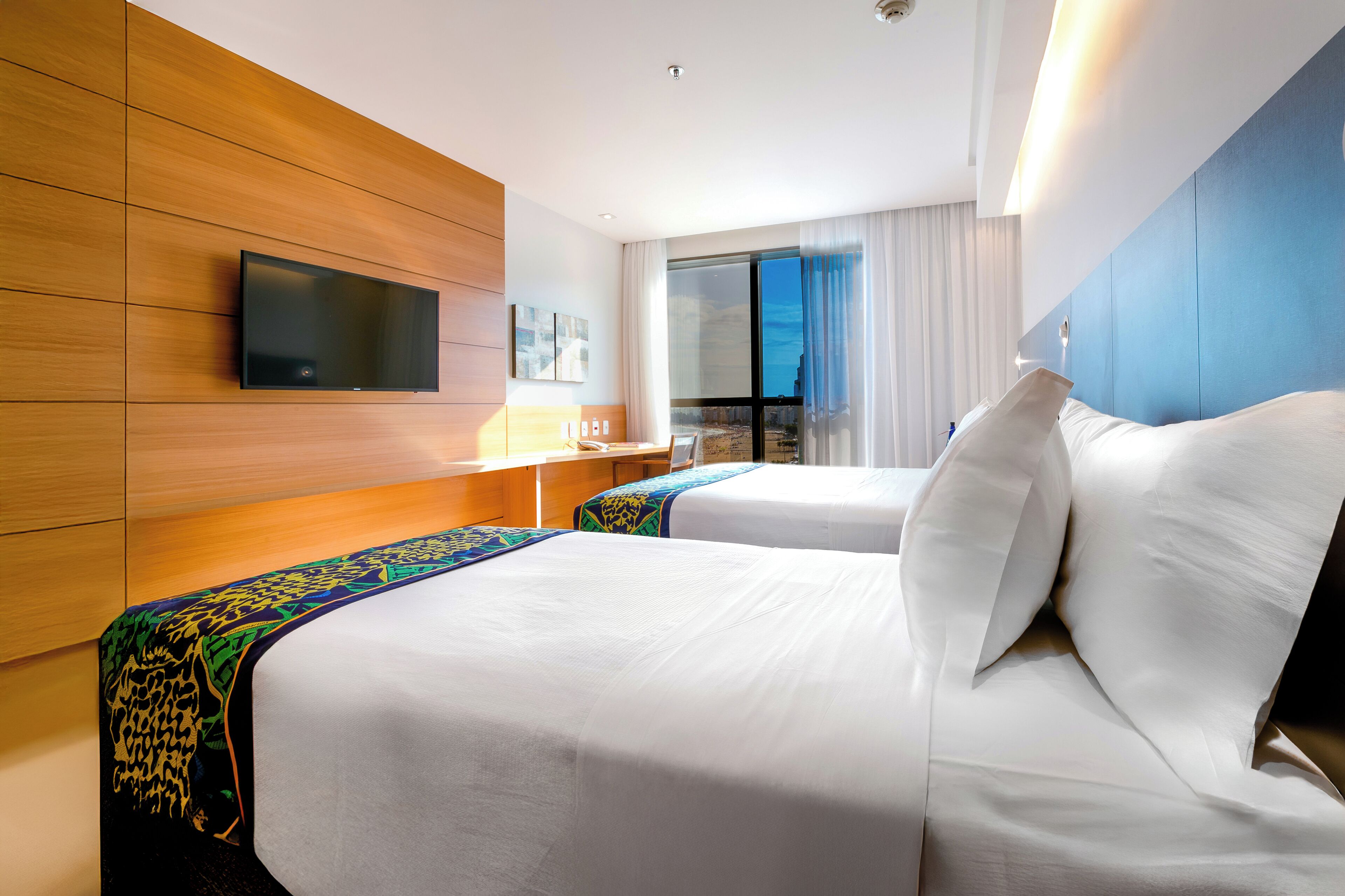 A hotel room with two beds, a TV mounted on the wall, a desk with a lamp, and a large window with curtains.