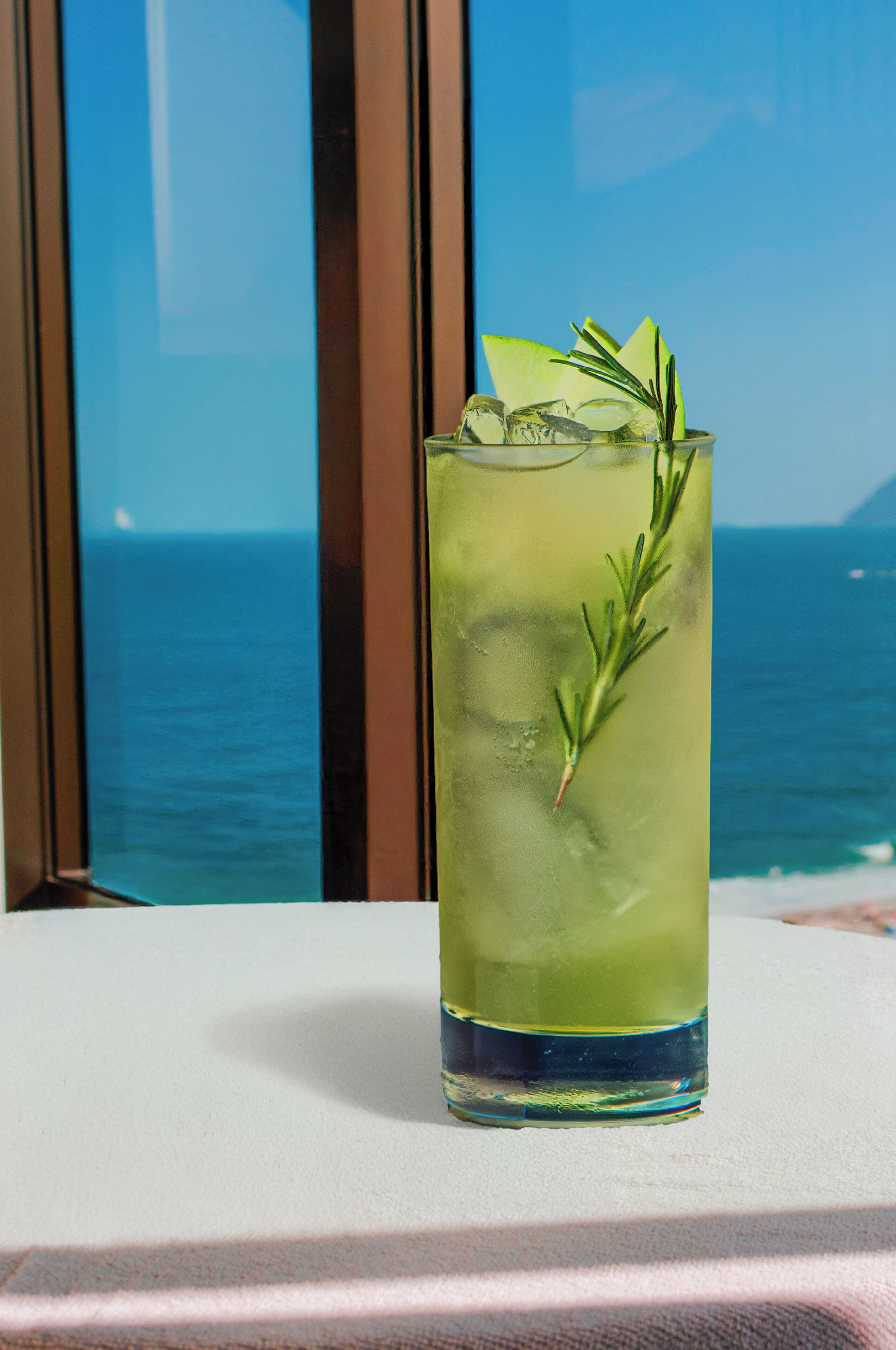 A tall glass with a green drink, ice, and a rosemary sprig, placed on a table with a view of the ocean through a window.