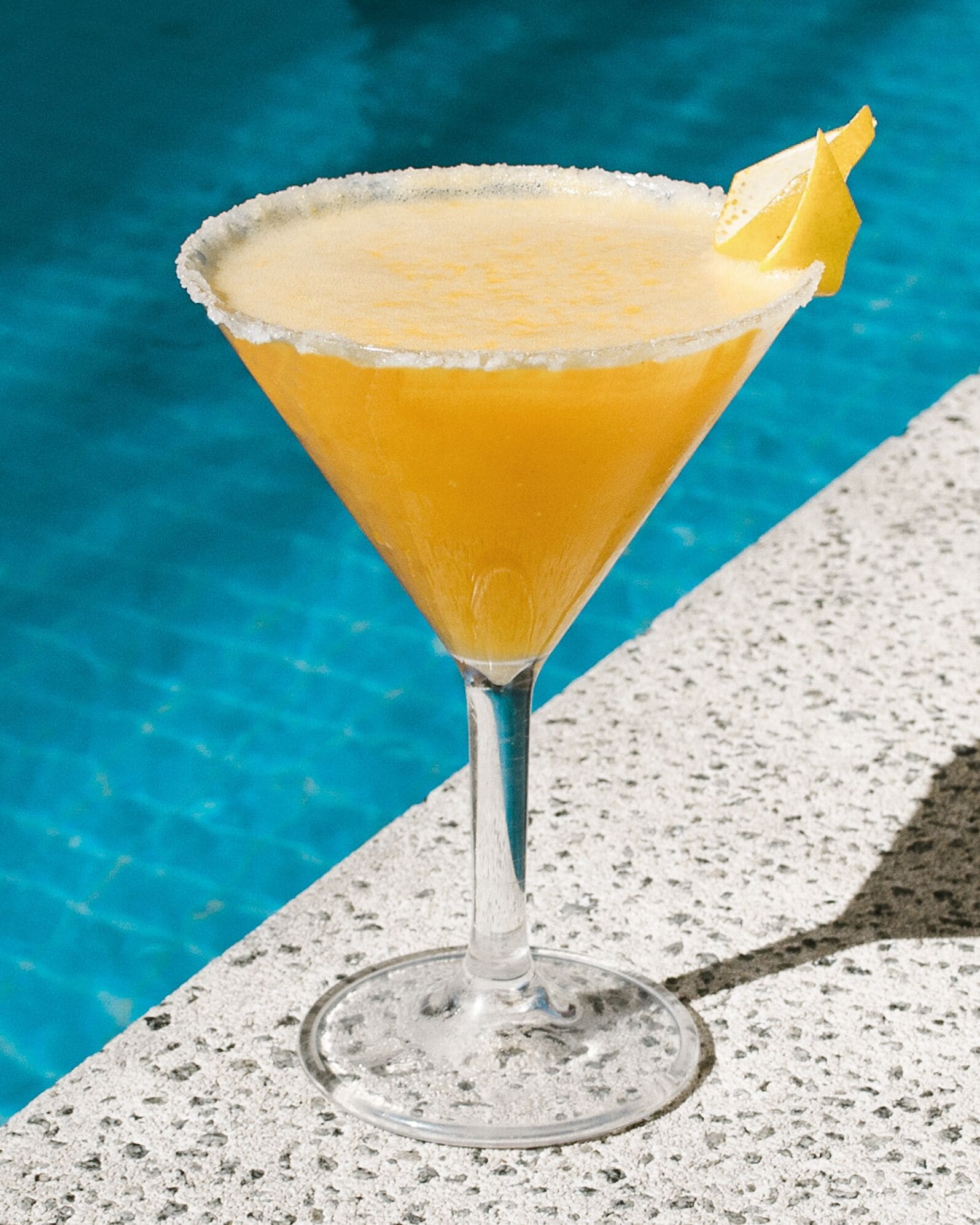 Um copo de cocktail com borda salgada e um toque de limão, apoiado em superfície de concreto perto de uma piscina.