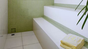 Un baño con paredes de azulejos verdes y blancos, un banco blanco y una pila de toallas amarillas.
