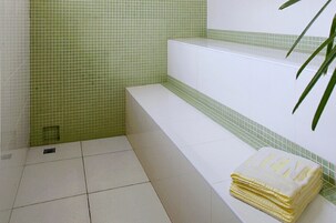 A bathroom with green and white tiled walls, a white bench, and a stack of yellow towels.