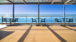 Ein sonnendurchfluteter Sitzbereich im Freien mit freiem Blick auf das Meer, ausgestattet mit Metalltischen und -stühlen sowie einer Glasüberdachung.