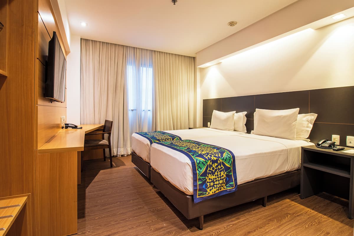 A hotel room with a bed, a desk, a chair, a TV mounted on the wall, and a window with curtains.