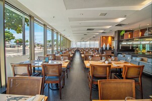 Um restaurante amplo com mesas e cadeiras compridas, grandes janelas com vista para a rua e uma área de cozinha visível à direita.