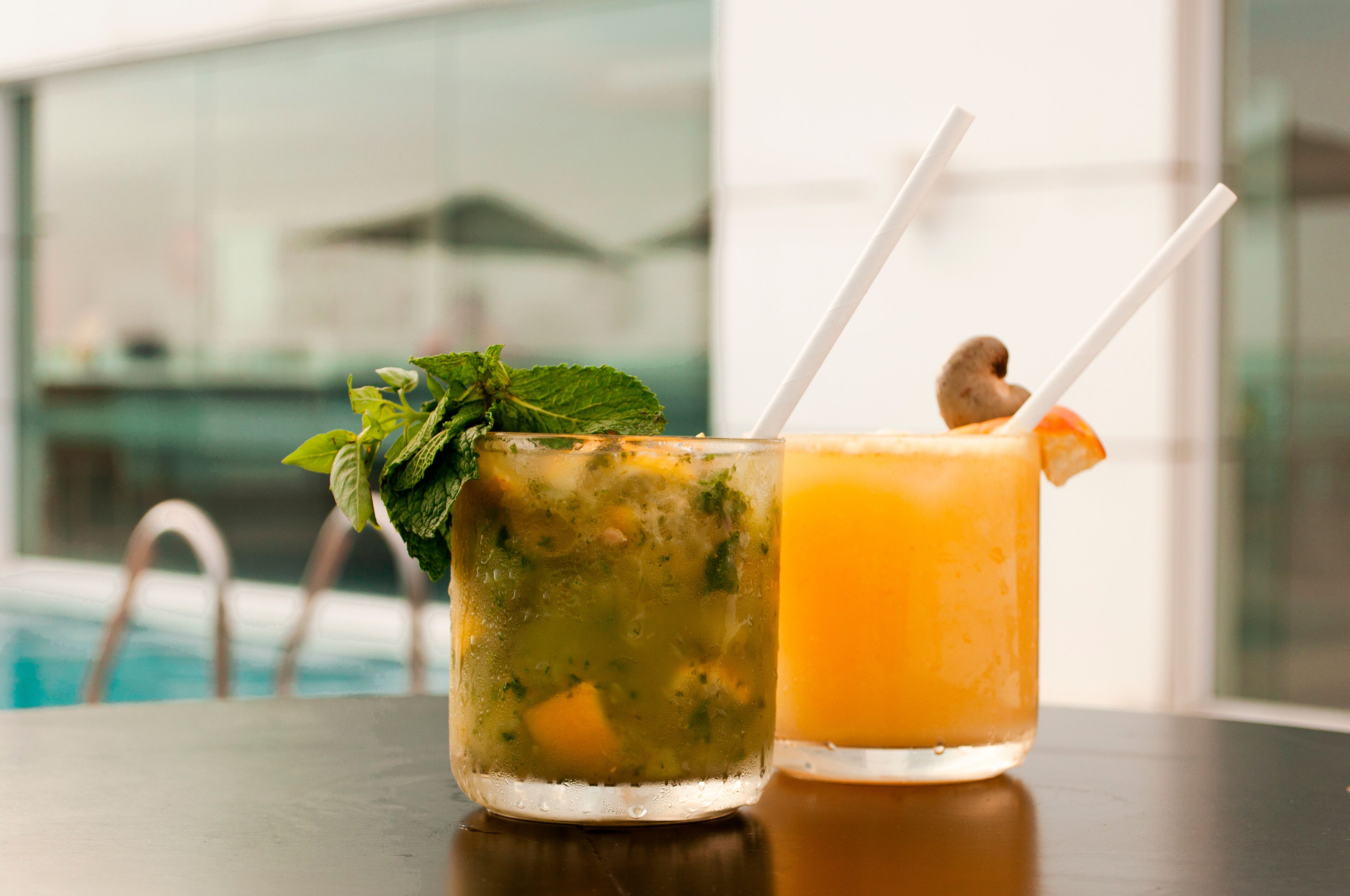 Zwei Getränke mit Trinkhalmen, eines mit Minze und das andere mit Orangenscheibe, auf einem Tisch in der Nähe eines Pools platziert.