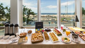 Uma mesa de buffet com diversos alimentos e vista para a praia através de grandes janelas.