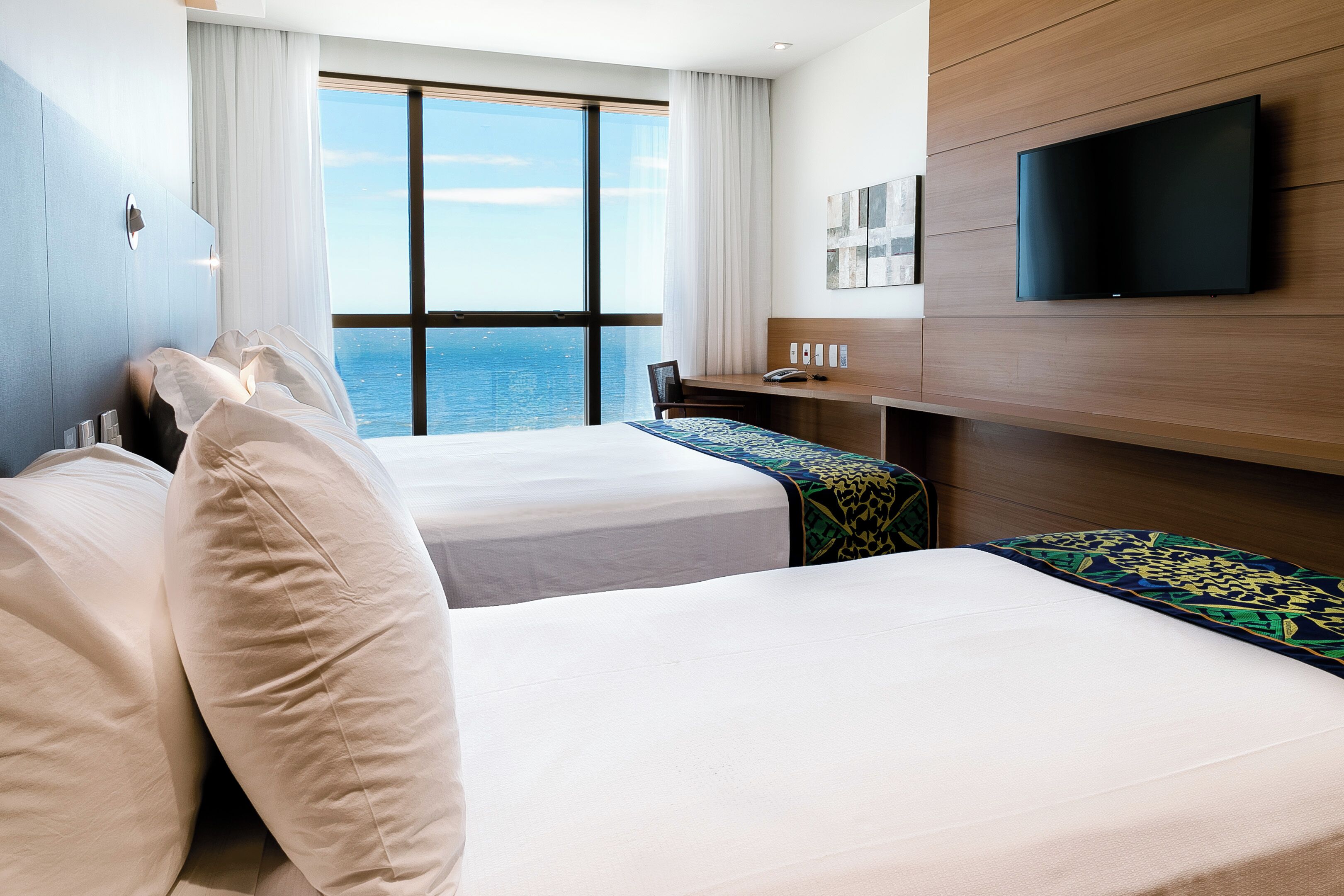 Habitación de hotel con dos camas, un escritorio, un televisor y un amplio ventanal con vistas al mar.