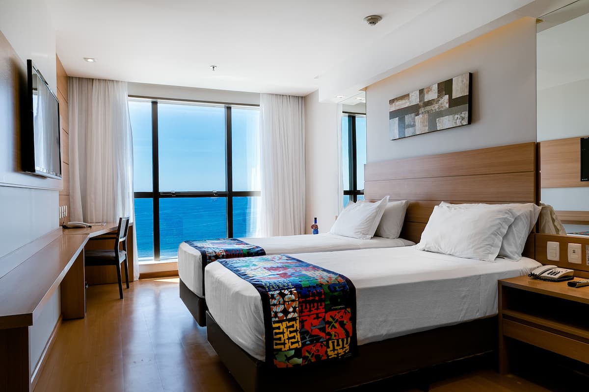A hotel room with two beds, a desk, and a large window offering a view of the ocean.