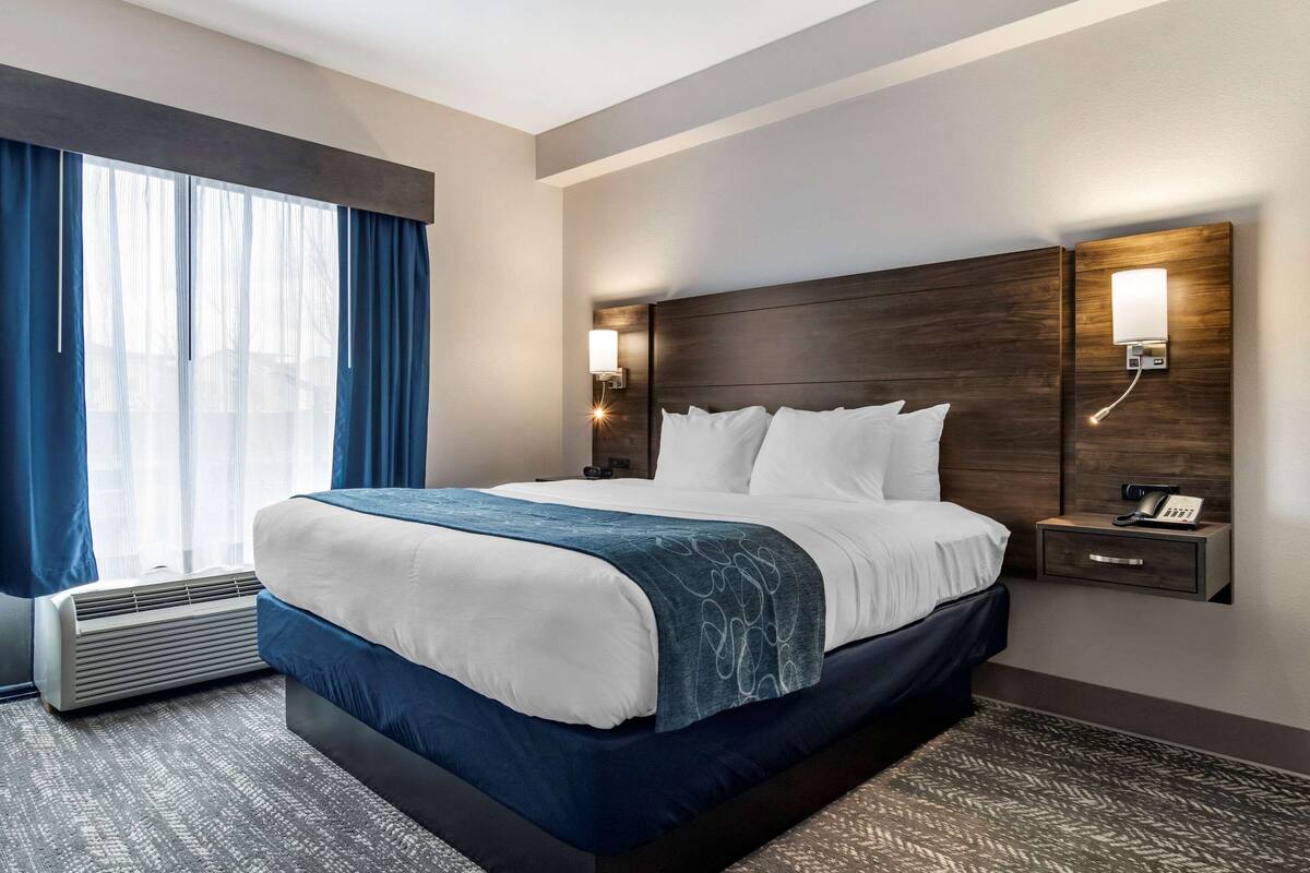Een hotelkamer met een groot bed, een nachtkastje met een telefoon, een wandlamp en een raam met gordijnen.