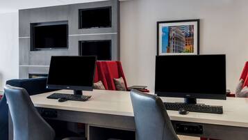 Un espacio de oficina moderno con dos escritorios, cada uno con monitor, teclado y ratón. Hay dos sillas, una con cojín rojo y otra con cojín gris. La habitación tiene una pared gris con estanterías empotradas y un cuadro enmarcado con una vista de la ciudad.