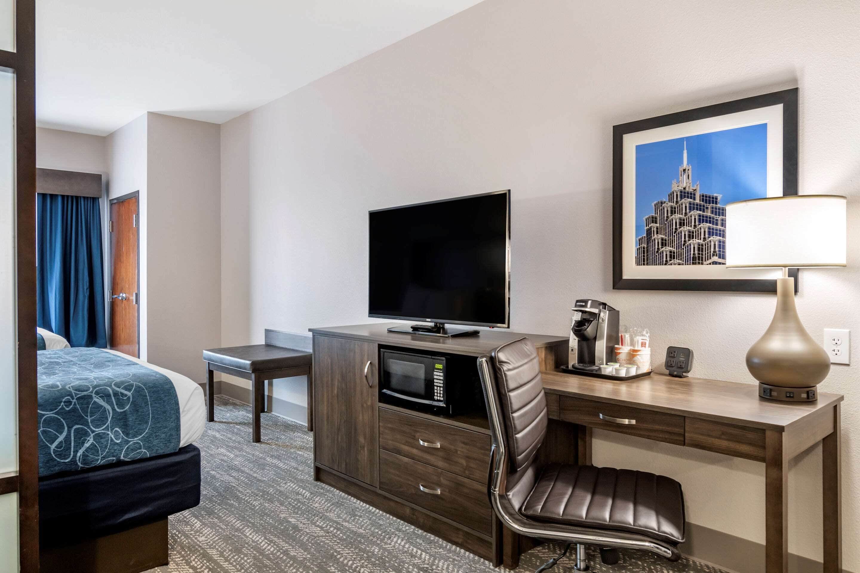 Una habitación de hotel con una cama, un escritorio con televisor de pantalla plana, cafetera, lámpara y un cuadro en la pared.