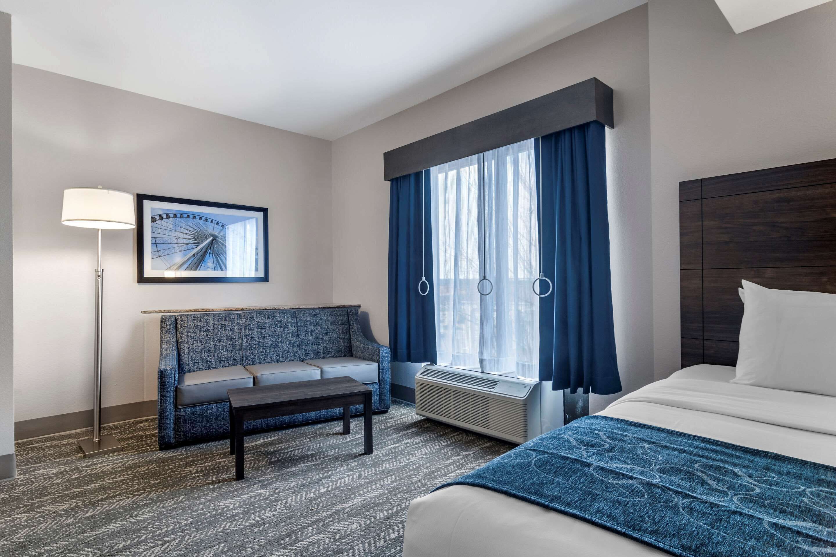 A hotel room with a bed, a sofa, a coffee table, a floor lamp, a picture on the wall, and a window with curtains.