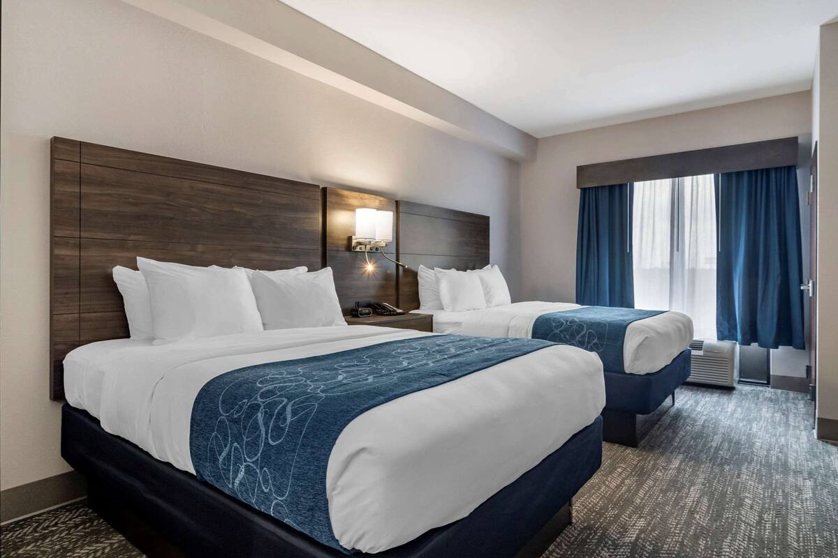 Een hotelkamer met twee bedden, een nachtkastje met een lamp en een groot raam met gordijnen.