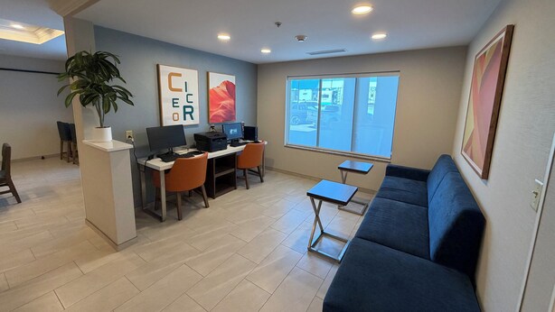 A modern office space with a desk, two chairs, a computer, a printer, a potted plant, and a large window with a view of the street.