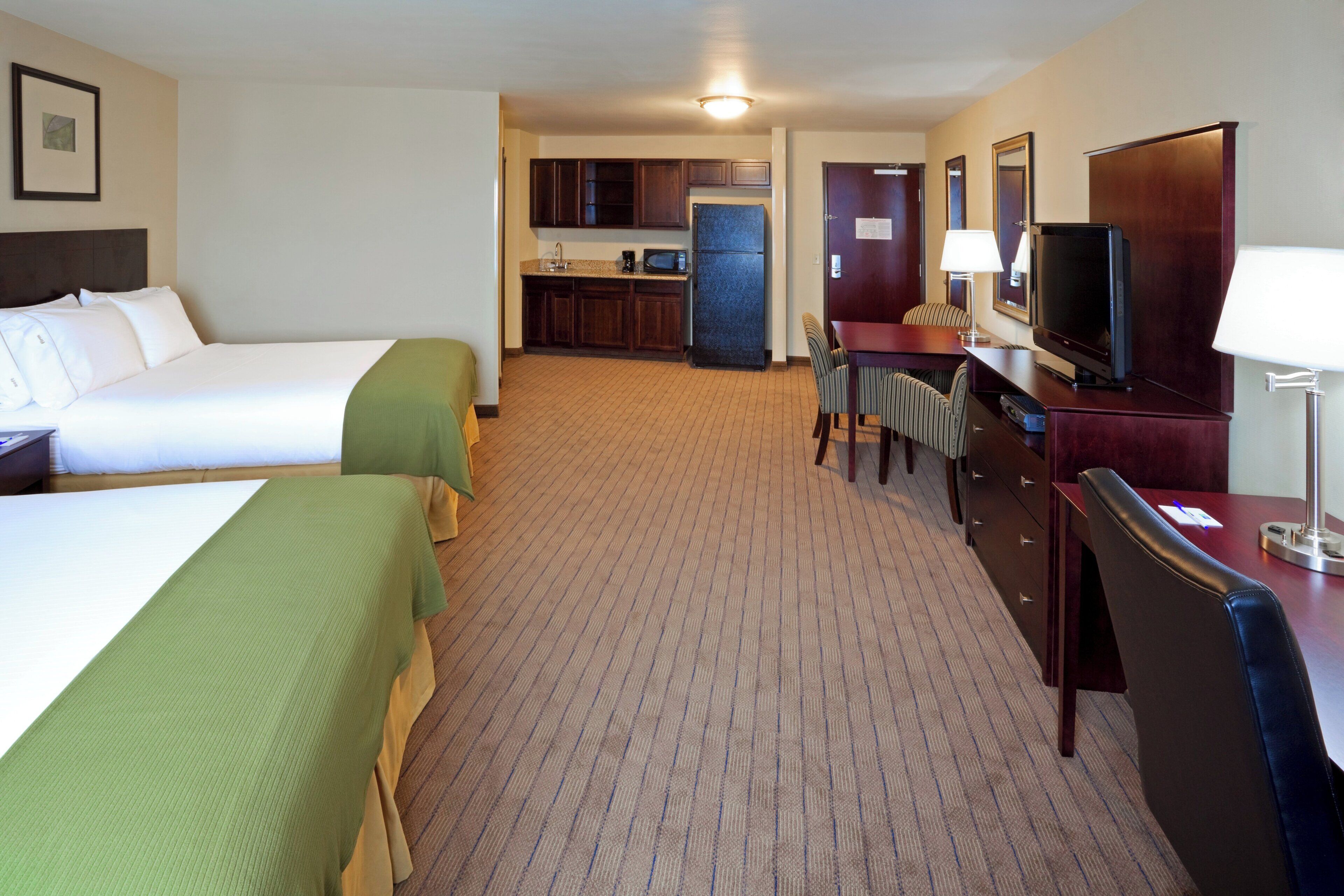 A hotel room with two beds, a kitchenette, a desk with a TV, and a chair.