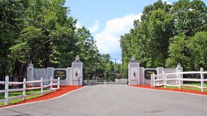 A gated entrance to a property with a white fence, a paved road, and lush green trees. - The Village At Pocono (Blakeslee)