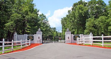 Una entrada protegida por una cerca blanca, un camino pavimentado y frondosos árboles verdes.