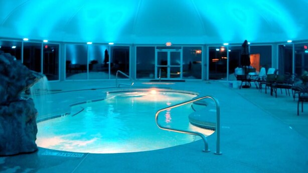 An indoor swimming pool with a glass barrier, surrounded by a covered area with seating and a view of the outside. - The Village At Pocono (Blakeslee)