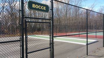 Una cerca de malla con un letrero que dice 'BOCCE' y una cancha de tenis al fondo.