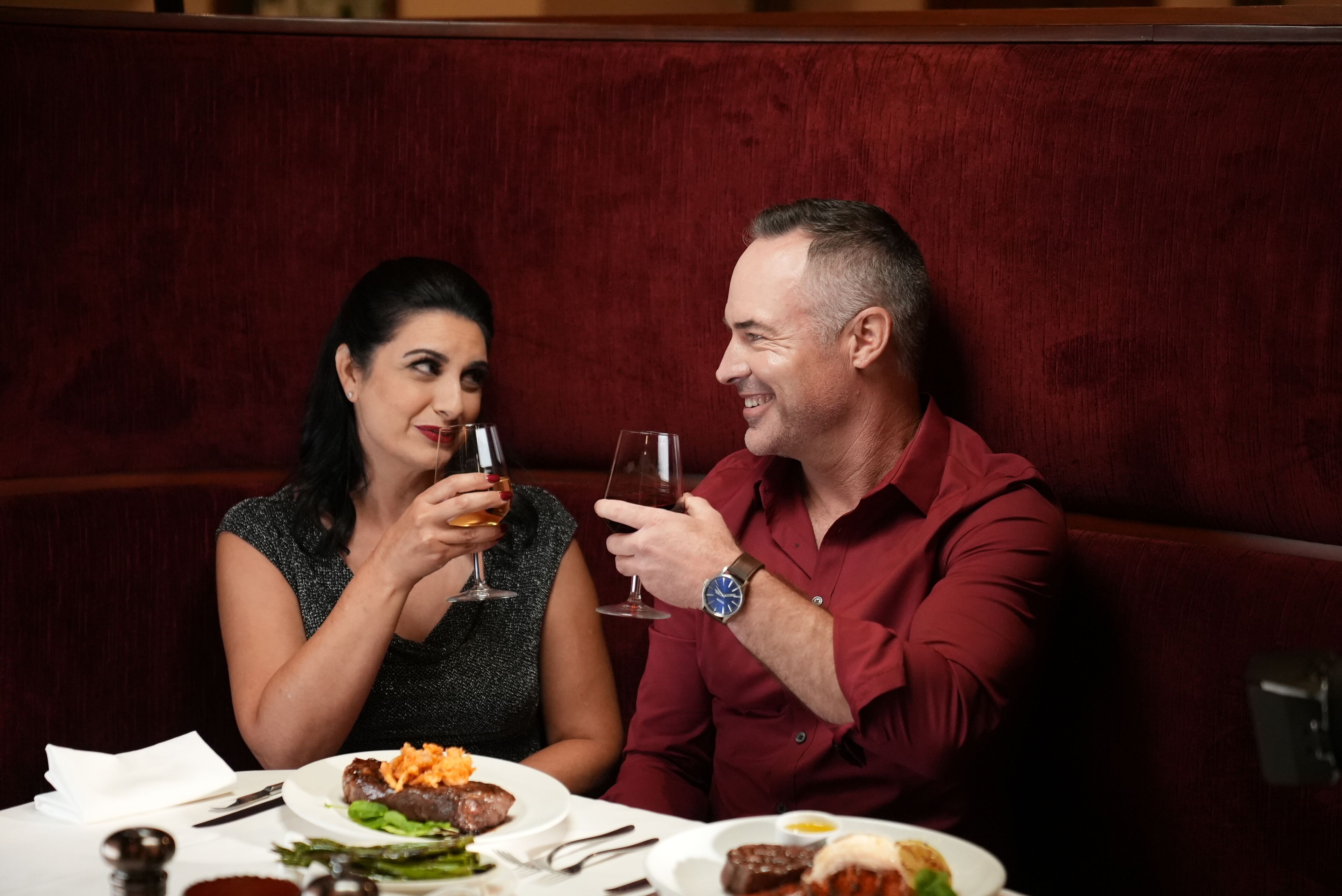 A couple seated at a table with a meal, in a room with red upholstered seating.