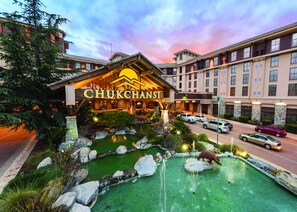 A hotel with a large sign reading "CHUKCHANSI", featuring a landscaped garden with a pond, a bear statue, and a parking area with several cars. - Chukchansi Gold Resort & Casino (Coarsegold)
