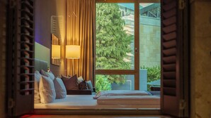 A hotel room with a bed, pillows, a nightstand, a lamp, and a window with a view of trees and buildings. - Qube Hotel Bergheim (Heidelberg)