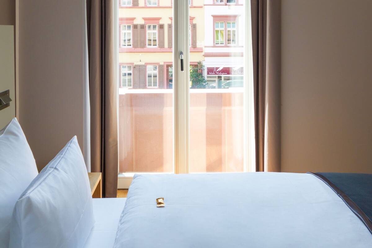 Habitación de hotel con cama, almohadas blancas, ventana con cortinas y vistas a edificios.