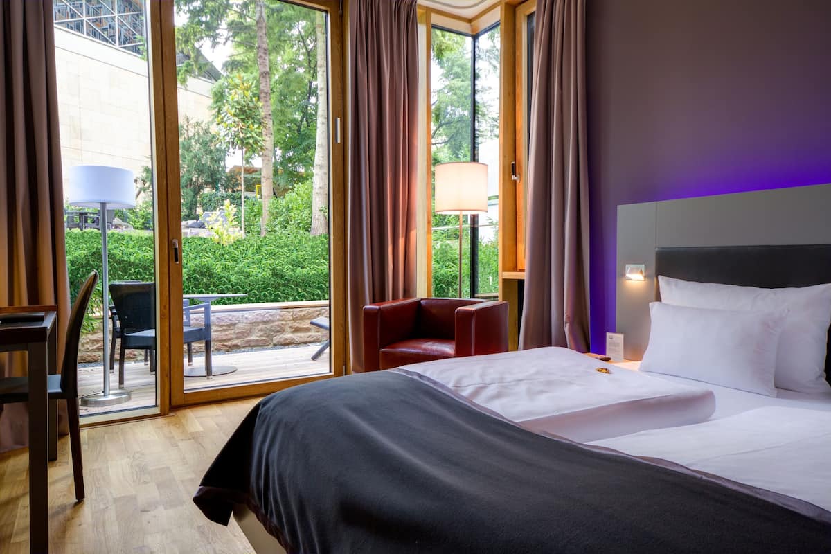 Habitación de hotel con cama, escritorio, silla y vistas a la vegetación a través de la puerta de vidrio.