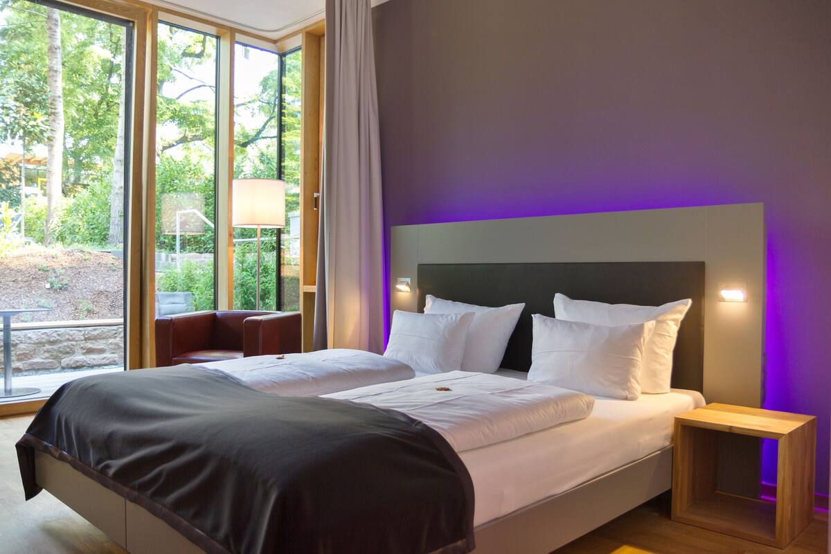 Una cama doble con cabecera oscura y ropa de cama blanca, una mesita de noche de madera y una ventana con vistas a la vegetación.