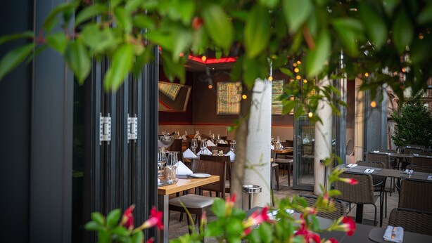 An outdoor dining area with tables and chairs, a glass wall, and a view of the interior. - Qube Hotel Bergheim (Heidelberg)