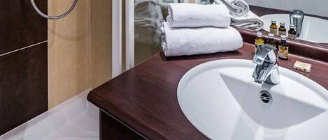 Une salle de bain avec un lavabo blanc, un miroir et une douche. On y trouve des serviettes et des articles de toilette sur le comptoir.