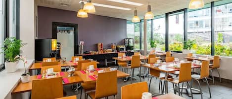 A spacious dining area with multiple tables and chairs, large windows offering a view of greenery outside, and pendant lighting.