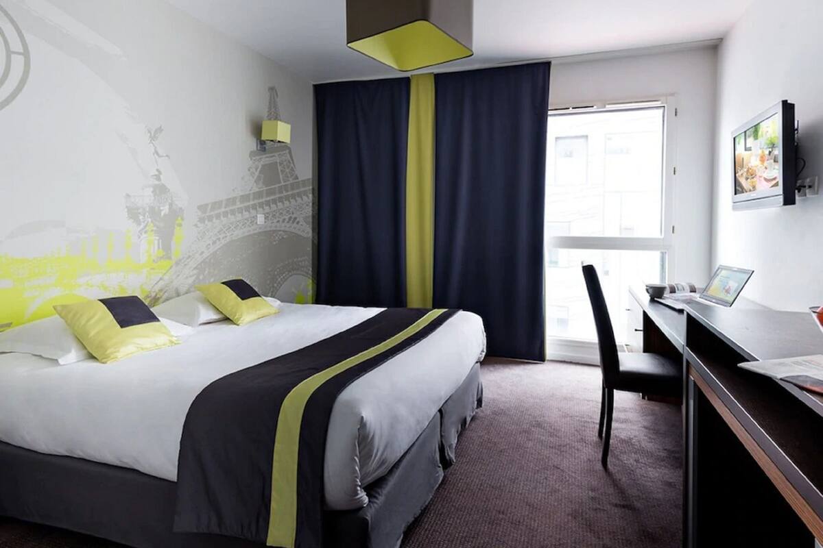 Ein Hotelzimmer mit Bett, Schreibtisch mit Laptop, Stuhl, großem Fenster mit Vorhängen und Wandgemälde.