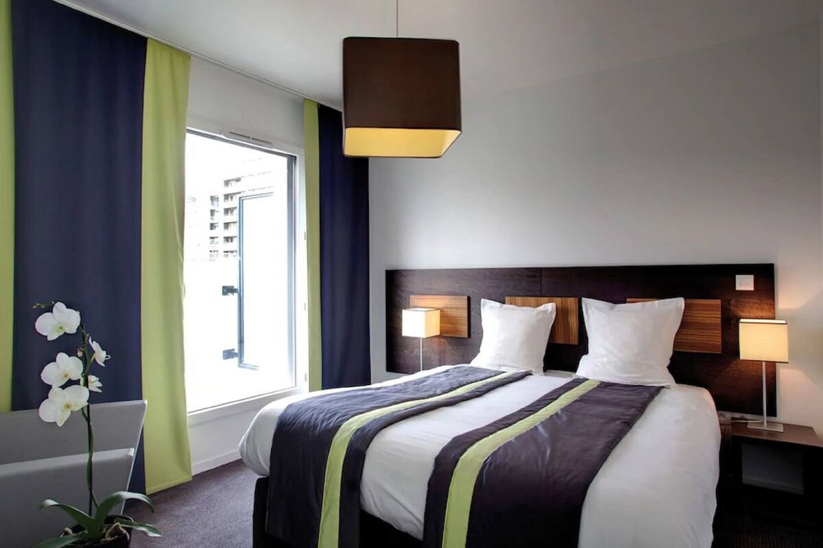 Ein Hotelzimmer mit einem großen Bett, einem Nachttisch mit Lampe, einem Fenster mit Vorhängen und einer Zimmerpflanze.