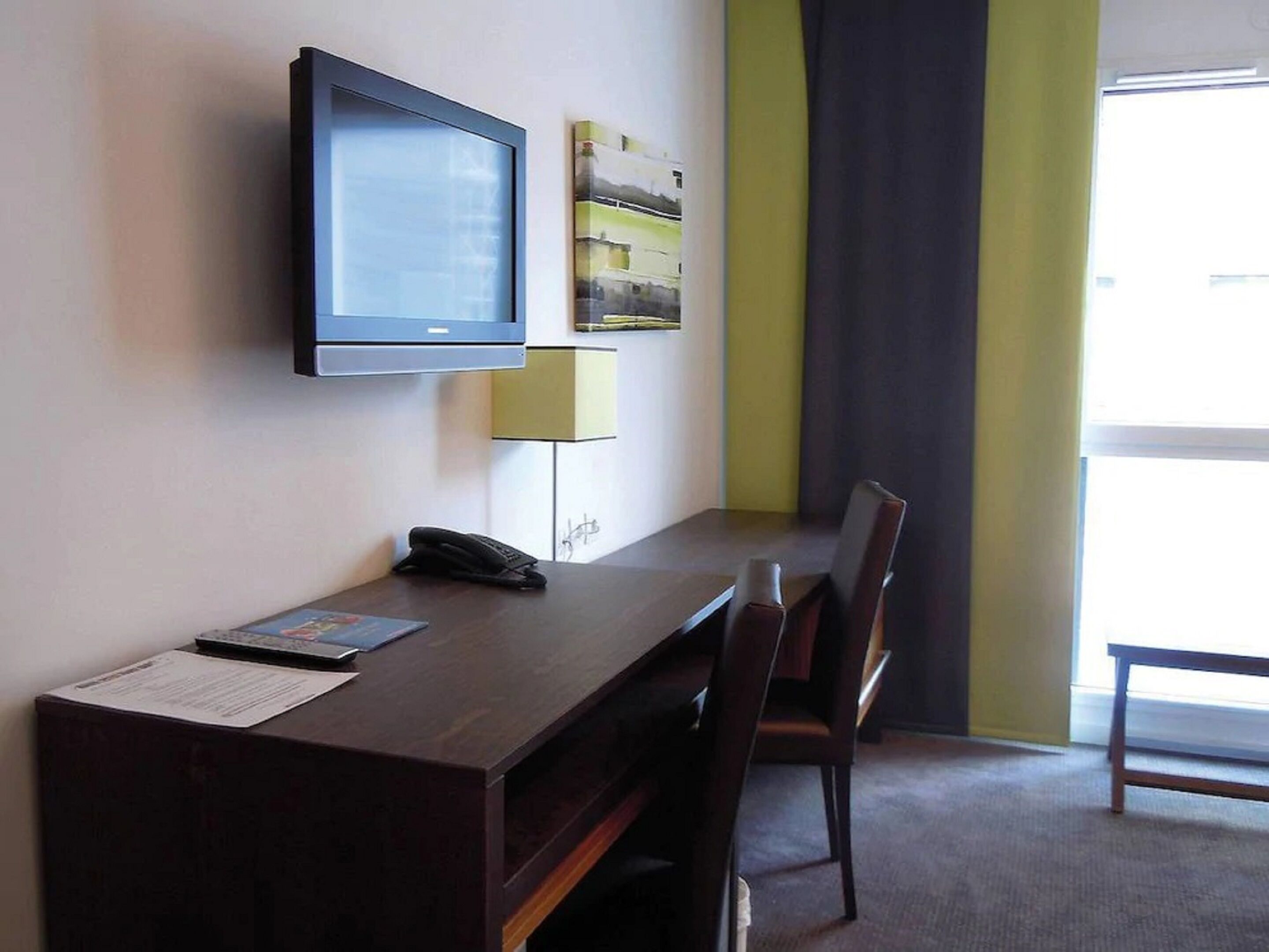 A hotel room with a desk, chair, telephone, and a television mounted on the wall.