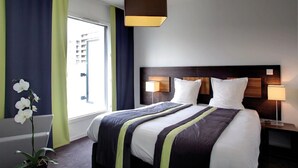 Ein Hotelzimmer mit einem großen Bett, einem Nachttisch mit Lampe, einem Fenster mit Vorhängen und einer Zimmerpflanze.