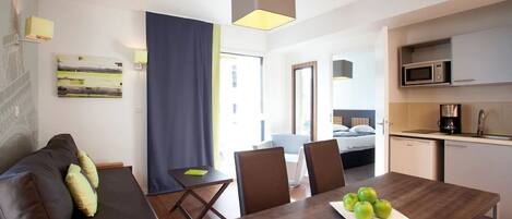 A modern hotel room with a wooden dining table, a sofa, and a kitchen area.