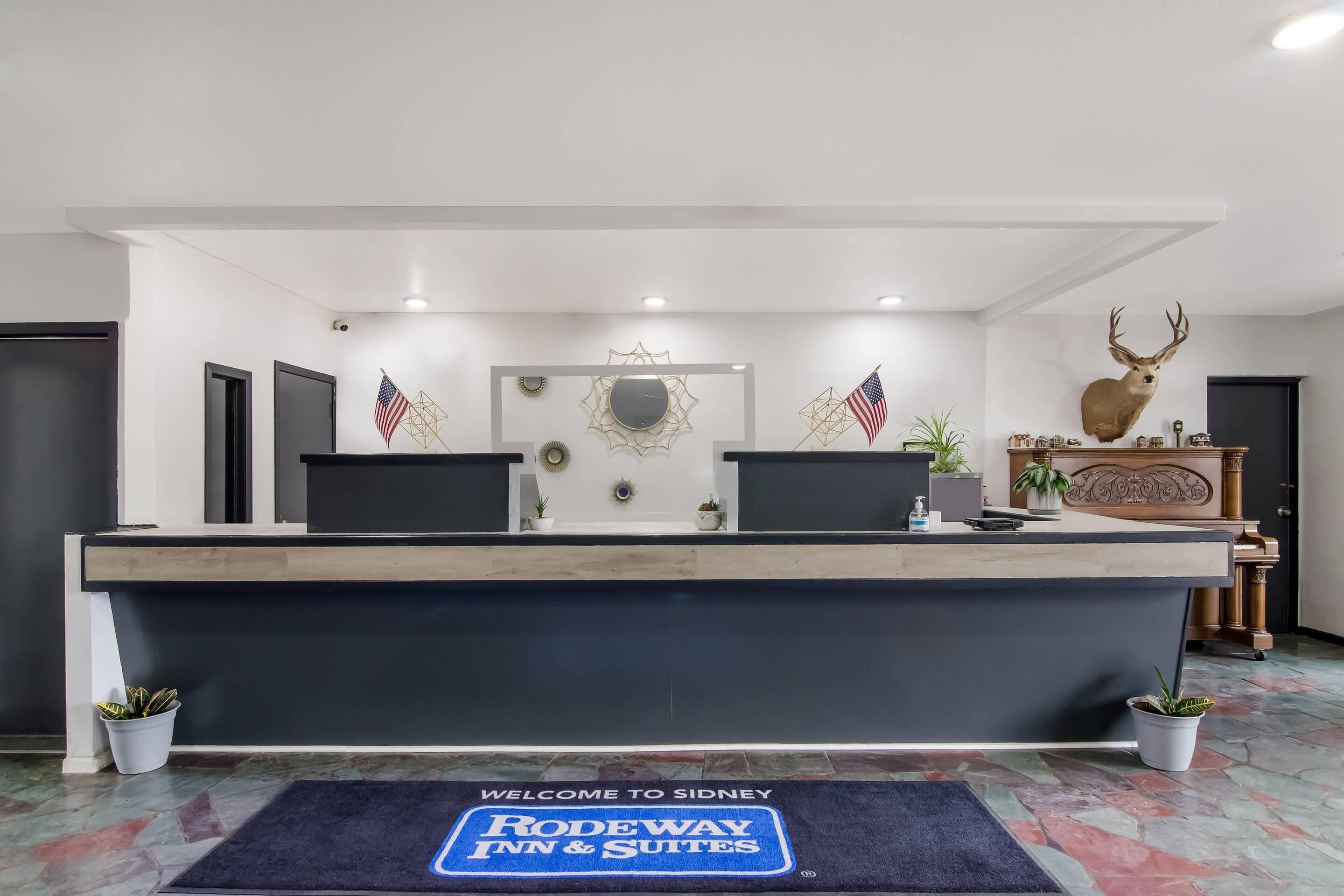 Tauleller de recepció a l'hotel Sidney Rodeway Inn & Suites, amb un estenedora de benvinguda, banderes americanes i un cap de cérvol penjat.