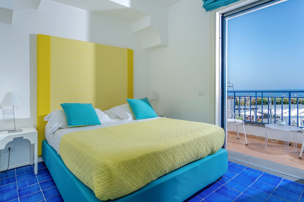 Habitación de hotel con cama, mesita de noche, lámpara y balcón con vista al mar.