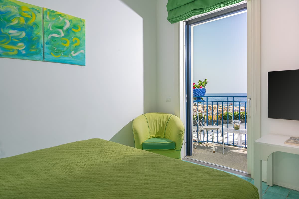 Una habitación con una cama, una silla verde, un televisor y un balcón con vistas al mar.