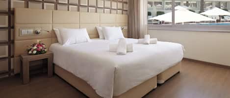 A hotel room with a large bed, a nightstand with a flower arrangement, and a view of the pool area through the window.