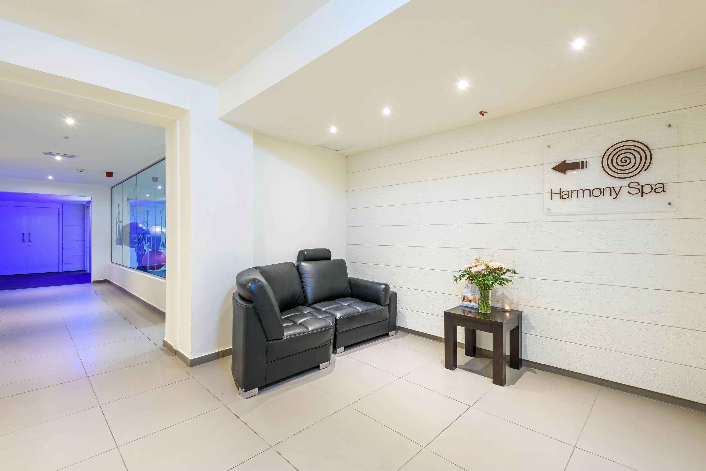Um hall de spa moderno com um sofá de couro preto, uma pequena mesa com flores e uma placa para Harmony Spa.