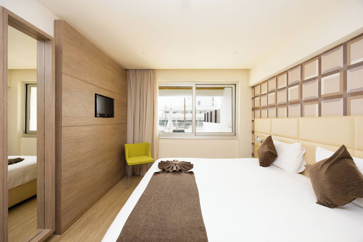 Een hotelkamer met een bed, een aan de muur bevestigde televisie, een raam met uitzicht op gebouwen en een gele stoel.