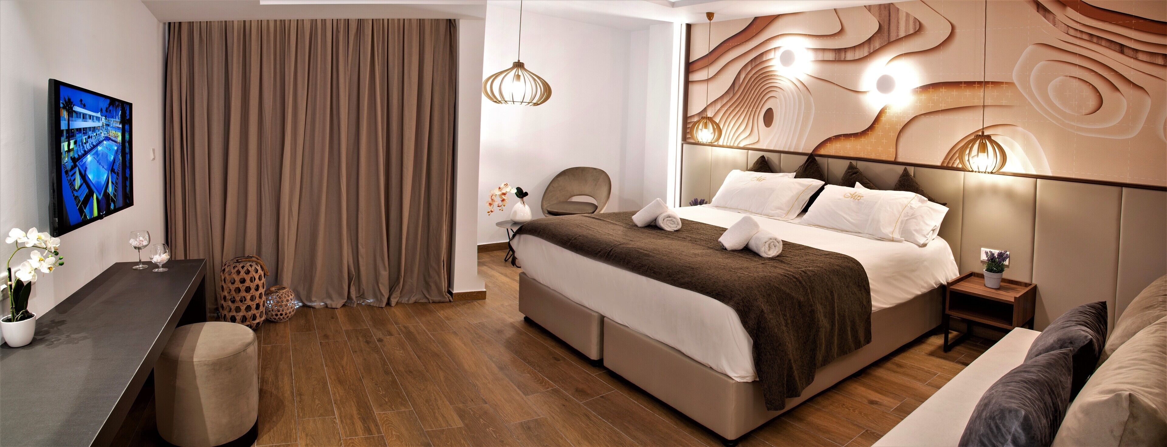 A modern hotel room with a large bed, a desk with a flat-screen TV, a chair, and a decorative wall.