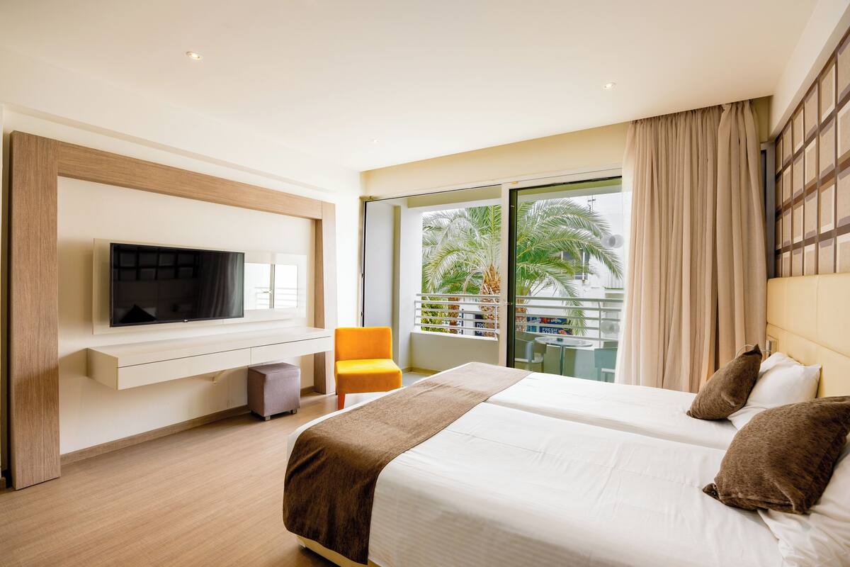Hotelkamer met een bed, een aan de muur bevestigde televisie, een stoel en een balkon met uitzicht op palmbomen.