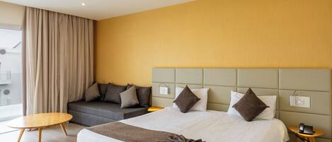 Una habitación de hotel moderna con una cama grande, un sofá, una mesita redonda y una mesita de noche con lámpara.