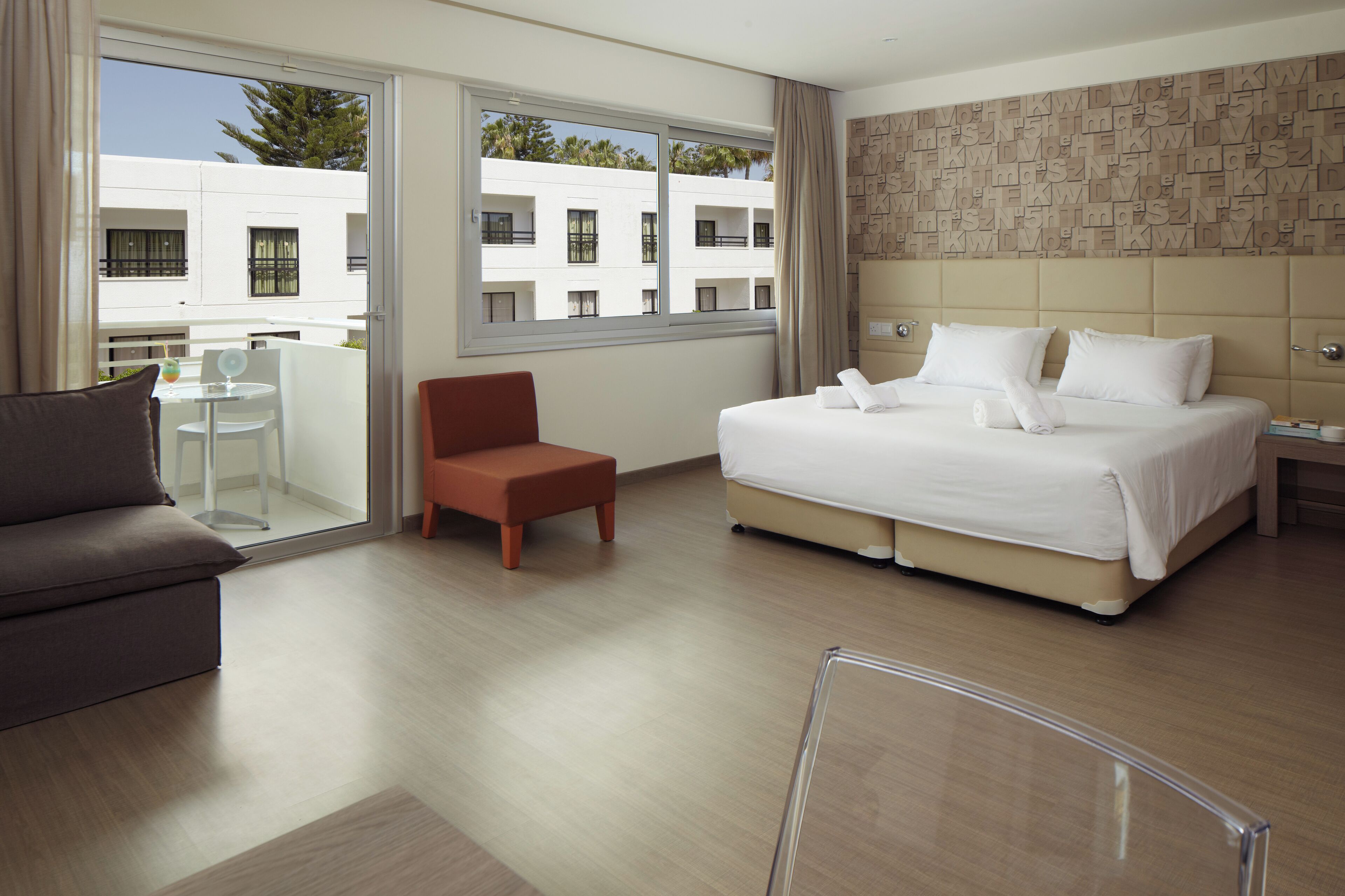 Une chambre d’hôtel avec un grand lit, un canapé, une petite table et une chaise. Elle dispose d’un balcon avec vue sur un immeuble et des arbres.