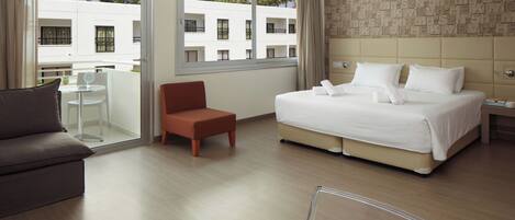 A hotel room with a large bed, a sofa, a small table, and a chair. There is a balcony with a view of a building and trees.