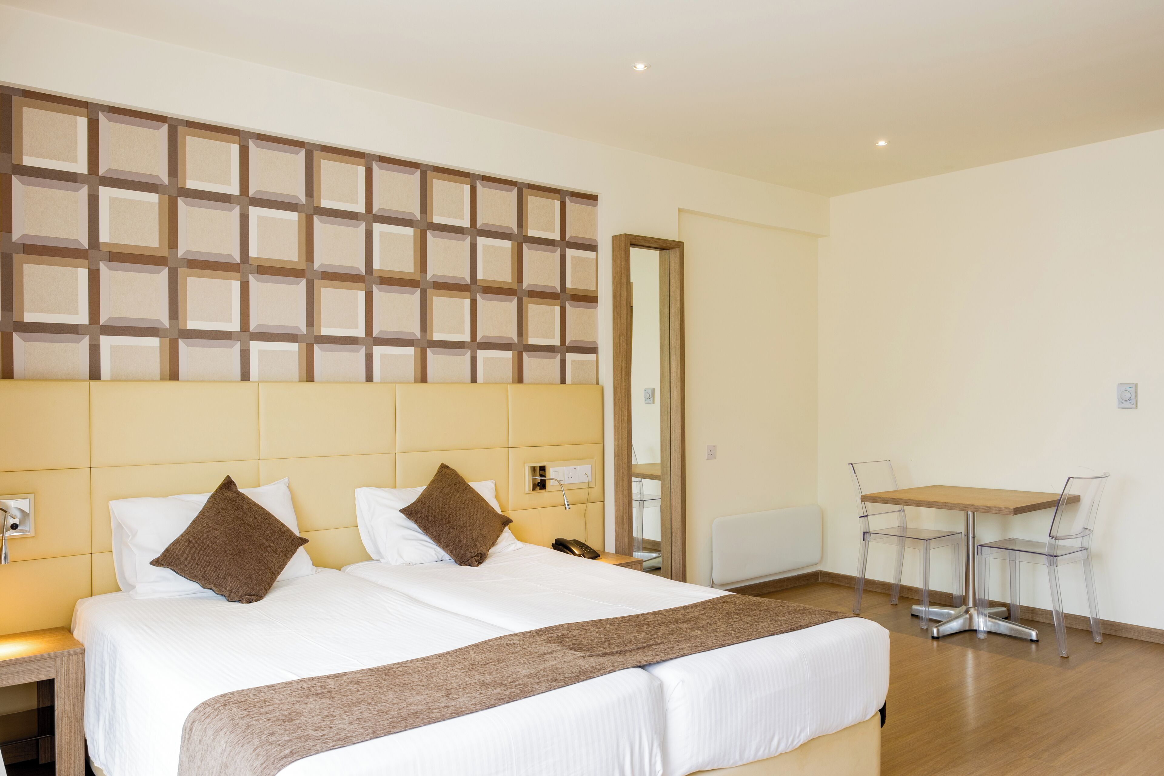 A hotel room with two beds, a wooden headboard, a small table with chairs, and a mirror.