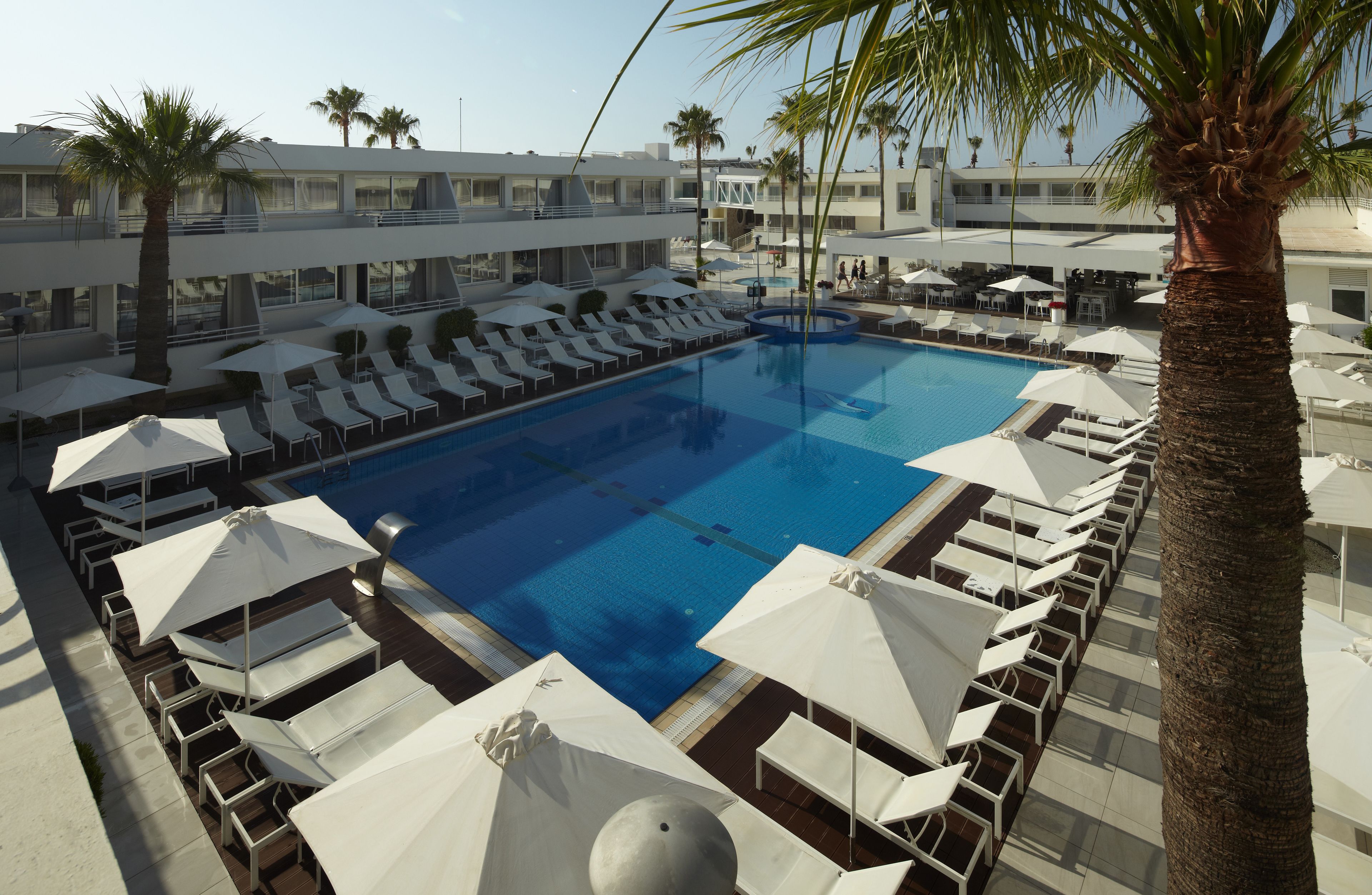 A pool area with sun loungers and umbrellas, surrounded by palm trees and a building with balconies.